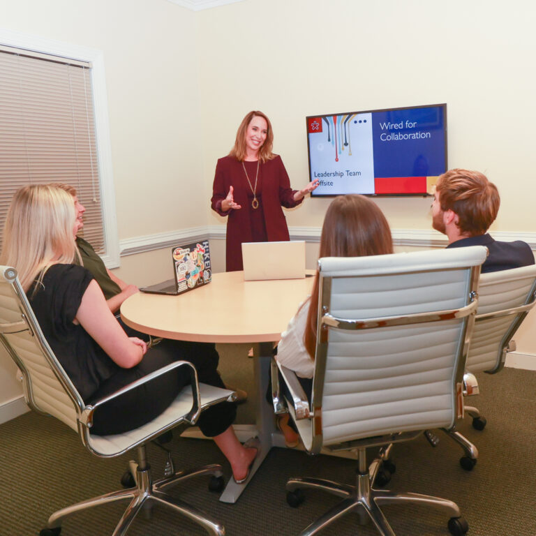 Image of Loretta wearing a burgundy dress and matching blazer standing in a small conference room next to a screen that reads "Wired for Connection." Her hands are in mid-gesture as she talks to people seated around the conference room table.