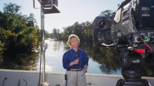 Old image of Loretta, standing on a bridge and holding a microphone; the shot shows a news camera and a studio light pointed at her.