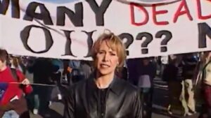 Old image of Loretta, wearing a black leather jacket and holding a microphone in front of a protest; a large banner behind her is not fully in frame, but visible text reads "Many dead ... oil???"