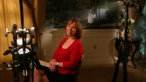 Old image of Loretta wearing a red sweater and black skirt, sitting in a news studio with cameras and lights around her.
