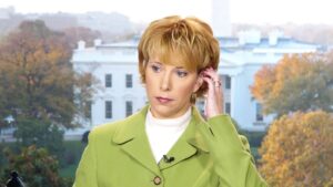 Image of Loretta wearing a green blazer with a lapel mic; she appears to be listening to something through an earpiece. The White House can be seen in the background.