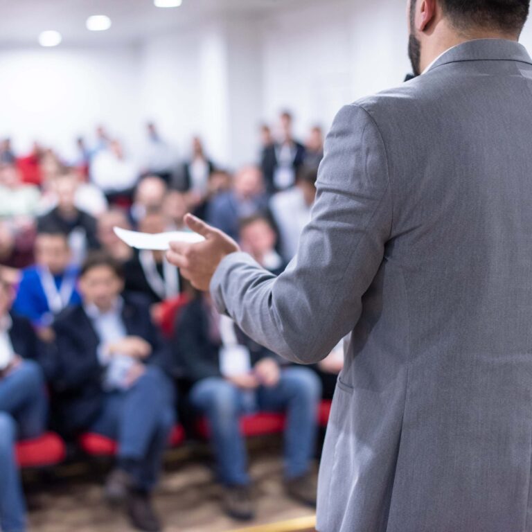 Photo of a man in a gray suit speaking to a medium-sized group.