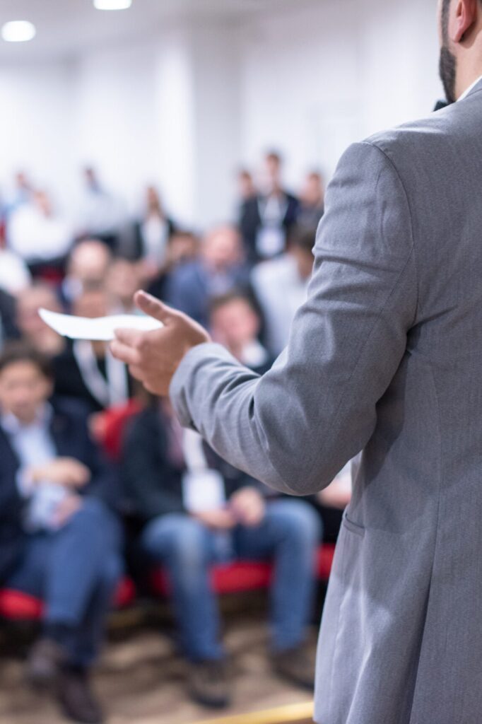 Photo from behind a man in a gray suit giving a presentation to a medium-sized group.