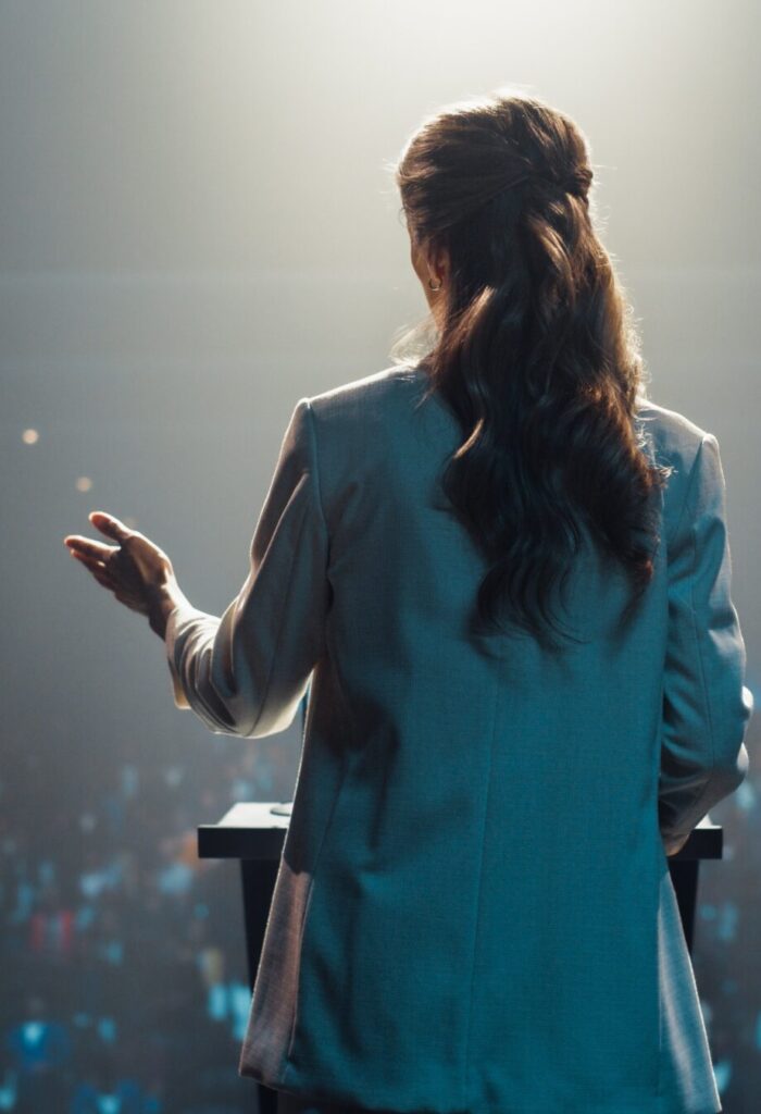 Photo from behind a woman on stage giving a speech to a large audience.