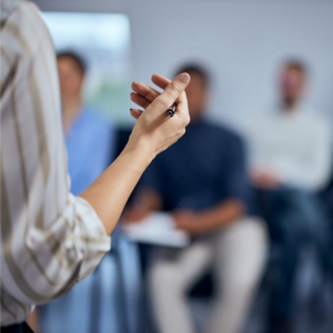 Photo from behind of the arm and shoulder of someone giving a presentation to a small team; the hand is holding a pen and seems to be mid-gesture, and a few people are visible taking notes behind it.